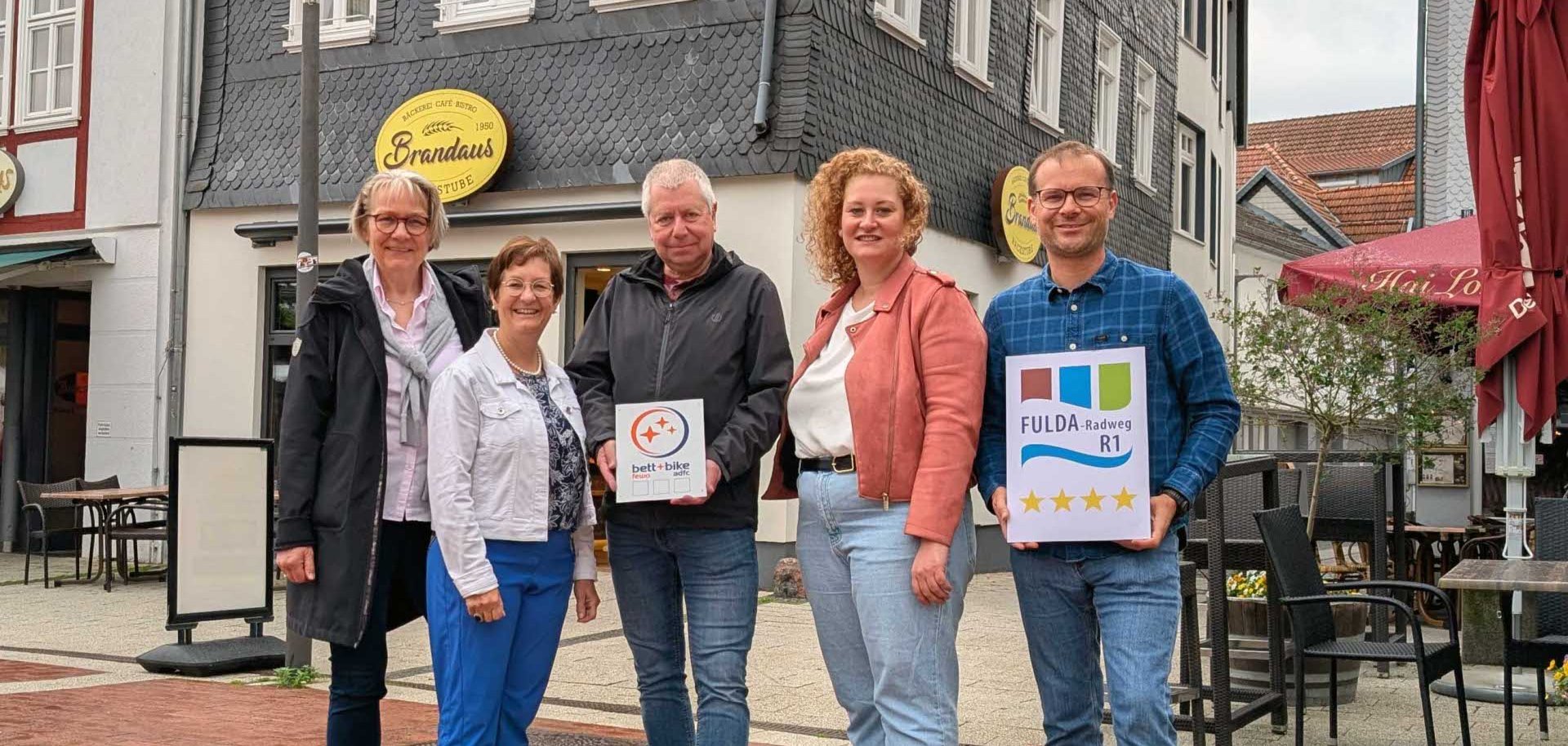 Apartments am Linggplatz- Bad Hersfeld: Beate Heiting Bett+Bike Hessen, Bürgermeisterin Anke Hofmann, Inhaber Matthias Zacharias, Franziska Rothenaicher (Fachbereich Kultur, Tourismus, Stadtmarketing Bad Hersfeld) Jörg Peters (Tourismusbeauftragter Landkreis Hersfeld-Rotenburg). Fünf Personen stehen vor dem Gebäude der „Apartments am Linggplatz“ in Bad Hersfeld. Zwei von ihnen halten Schilder zur Bett+Bike- und Fulda-Radweg-Zertifizierung.