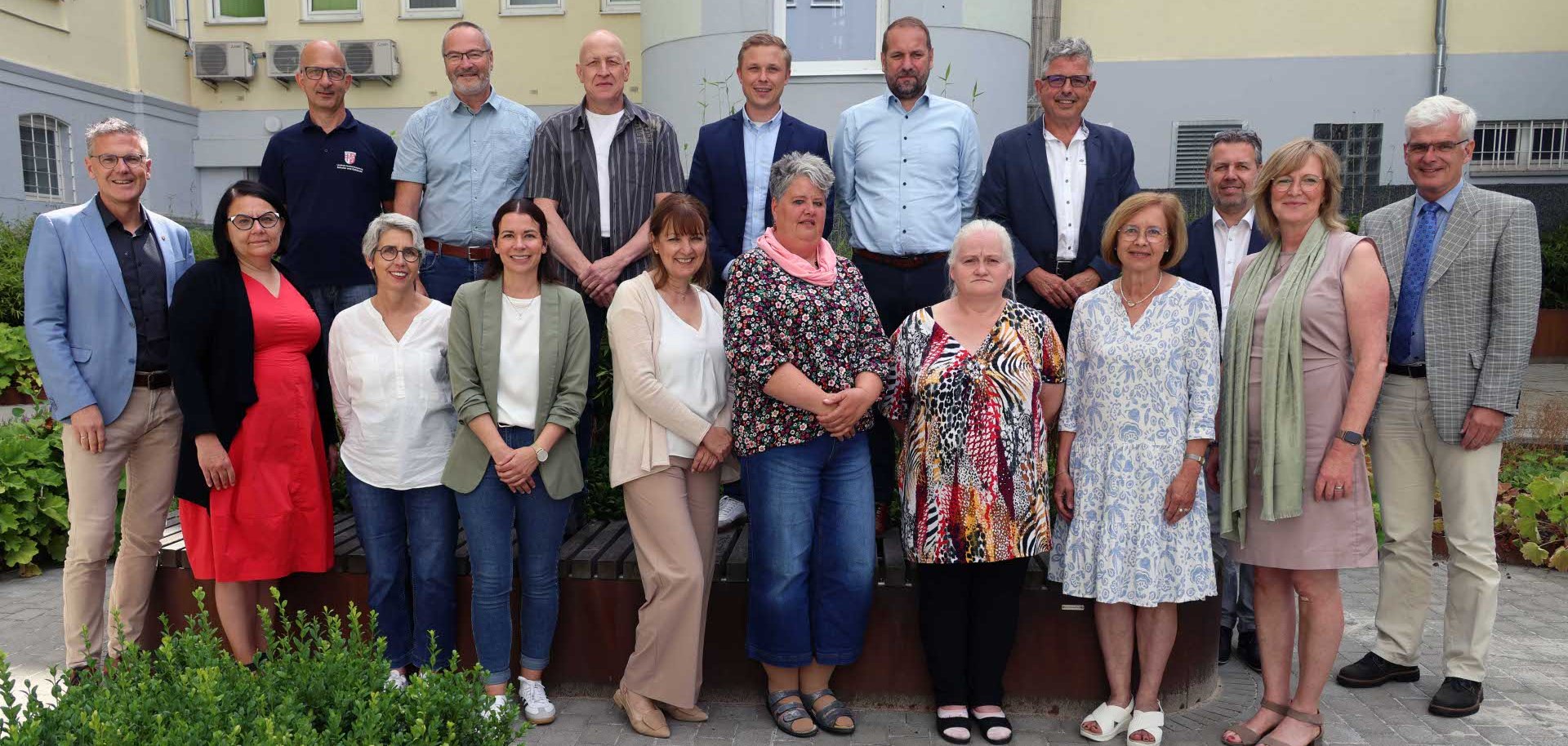 Die Gruppe besteht aus Männern und Frauen unterschiedlichen Alters, die in zwei Reihen posieren – die vordere Reihe steht auf dem Boden, die hintere leicht erhöht auf einem Holzpodest. Alle blicken lächelnd in die Kamera. Die Kleidung ist überwiegend sommerlich und leger bis semi-formell. Im Hintergrund sind Fenster, eine Tür, Klimaanlagen und Pflanzenkübel zu sehen. Das Foto scheint im Innenhof oder Eingangsbereich eines Amts- oder Schulgebäudes aufgenommen worden zu sein. Die Stimmung wirkt freundlich und professionell.