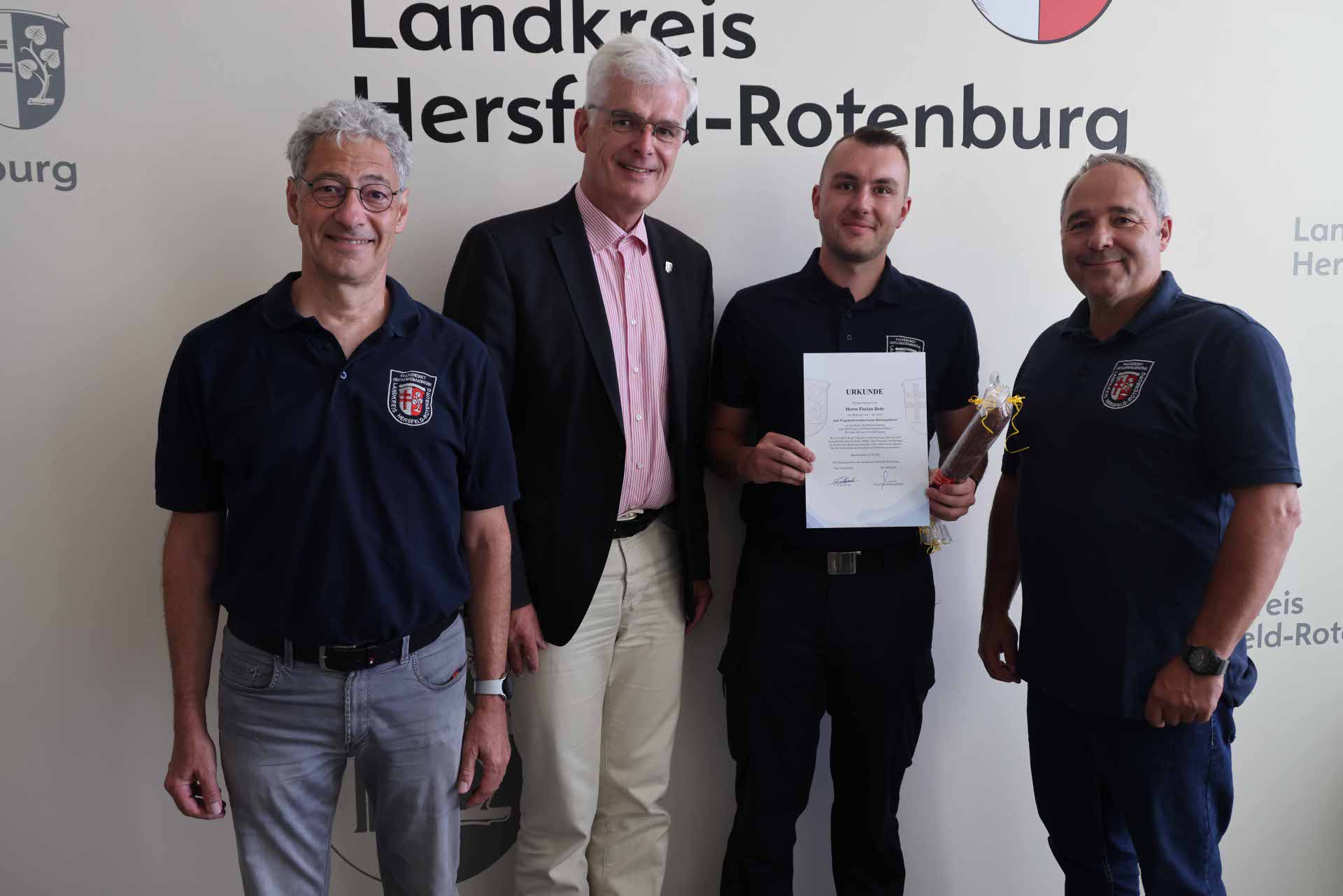Vier Männer stehen vor einer Wand mit dem Schriftzug „Landkreis Hersfeld-Rotenburg“. Einer von ihnen hält eine Urkunde und ein kleines Geschenk in der Hand.