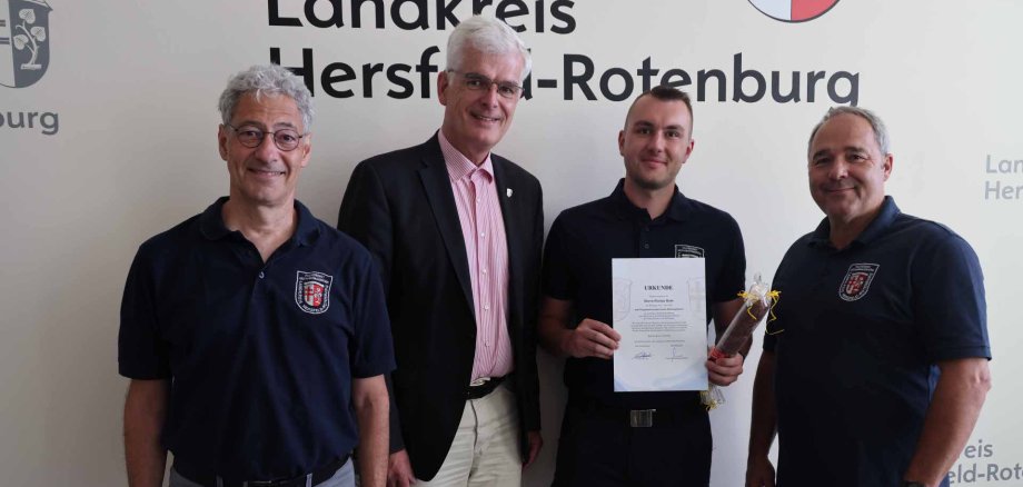 Vier Männer stehen vor einer Wand mit dem Schriftzug „Landkreis Hersfeld-Rotenburg“. Einer von ihnen hält eine Urkunde und ein kleines Geschenk in der Hand.