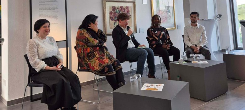 Fünf Personen sitzen auf einem Podium in einer Galerie. Im Hintergrund sind Kunstwerke und eine Leinwand mit Informationen zur Podiumsdiskussion zu sehen. Thema ist Migration und gesellschaftliches Miteinander.