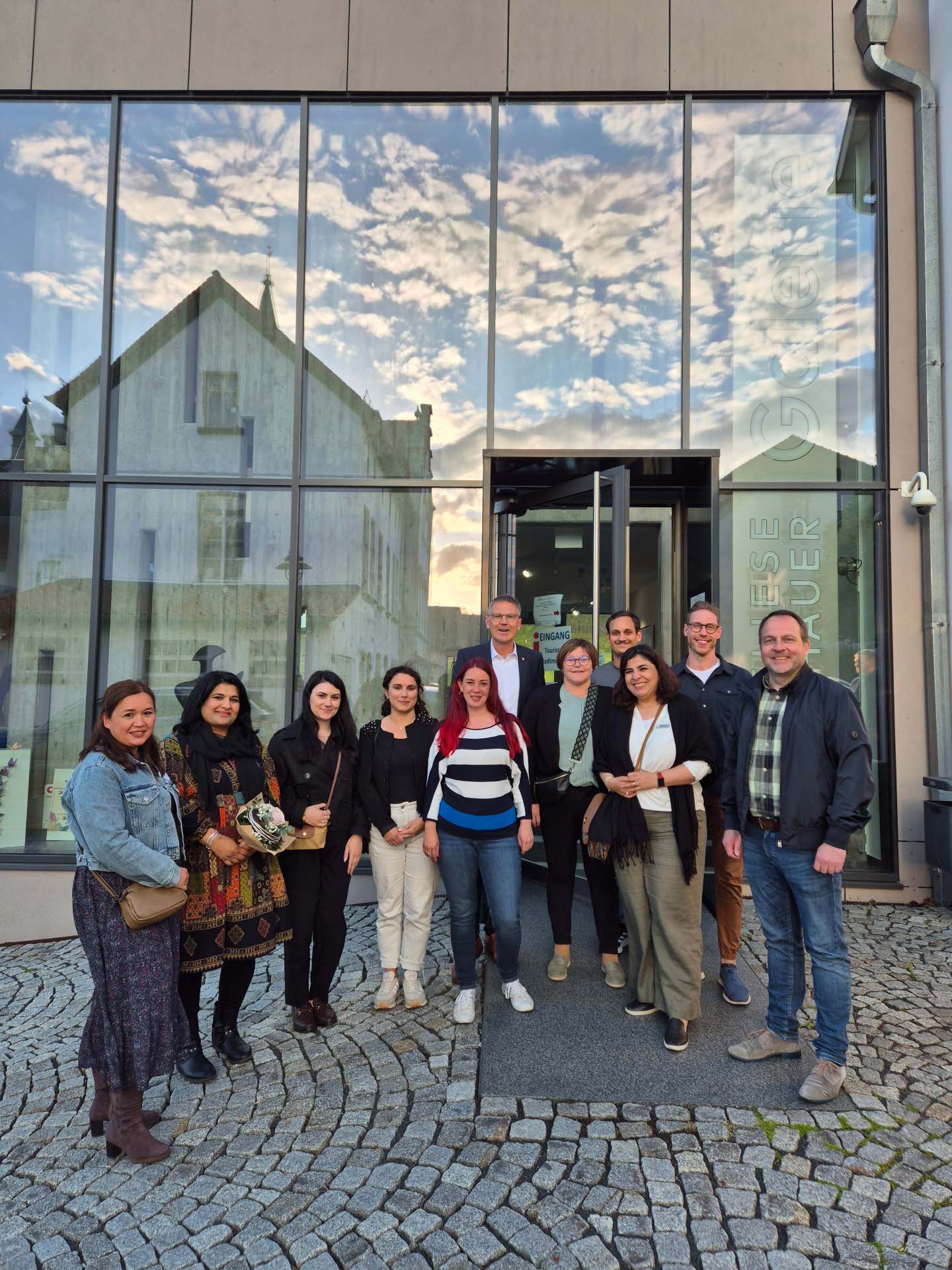 Gruppe von Teilnehmenden steht vor dem einem Gebäude und blickt lächelnd in die Kamera. Die Glasfassade spiegelt den Himmel mit Wolken wider.