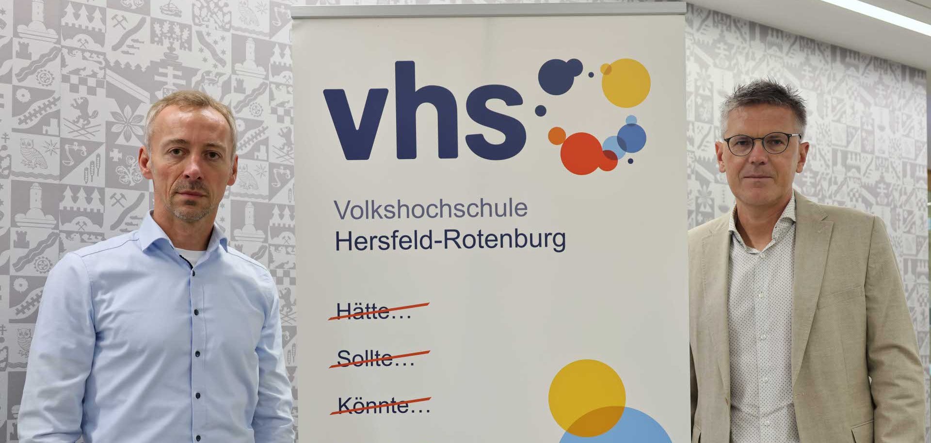 Zwei Männer stehen links und rechts neben einem Roll-Up der Volkshochschule Hersfeld-Rotenburg. Auf dem Roll-Up ist das vhs-Logo zu sehen sowie der motivierende Slogan: „Hätte… Sollte… Könnte… Würde… – Machen!“ Die durchgestrichenen Begriffe symbolisieren Entschlossenheit und Tatkraft. Im Hintergrund ist eine graue Wand mit stilisierten regionalen Motiven sichtbar.