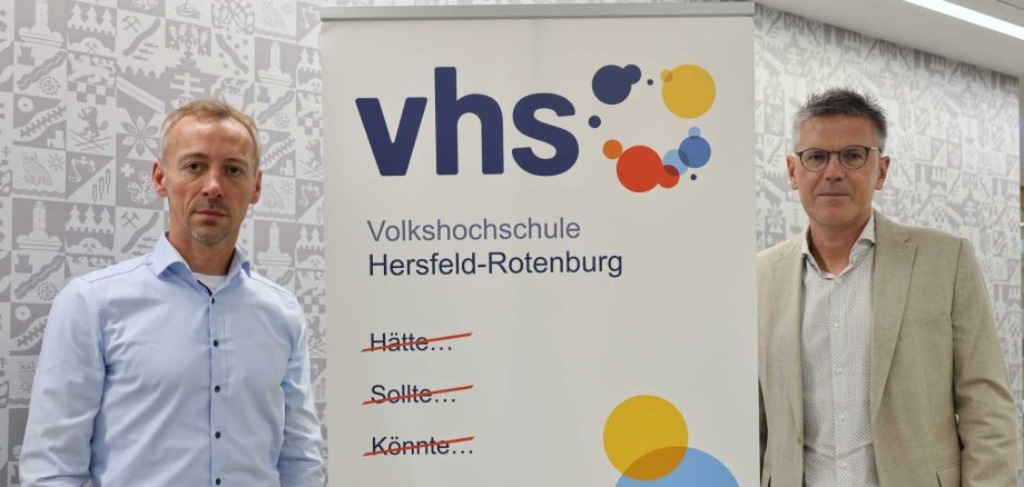 Zwei Männer stehen links und rechts neben einem Roll-Up der Volkshochschule Hersfeld-Rotenburg. Auf dem Roll-Up ist das vhs-Logo zu sehen sowie der motivierende Slogan: „Hätte… Sollte… Könnte… Würde… – Machen!“ Die durchgestrichenen Begriffe symbolisieren Entschlossenheit und Tatkraft. Im Hintergrund ist eine graue Wand mit stilisierten regionalen Motiven sichtbar.