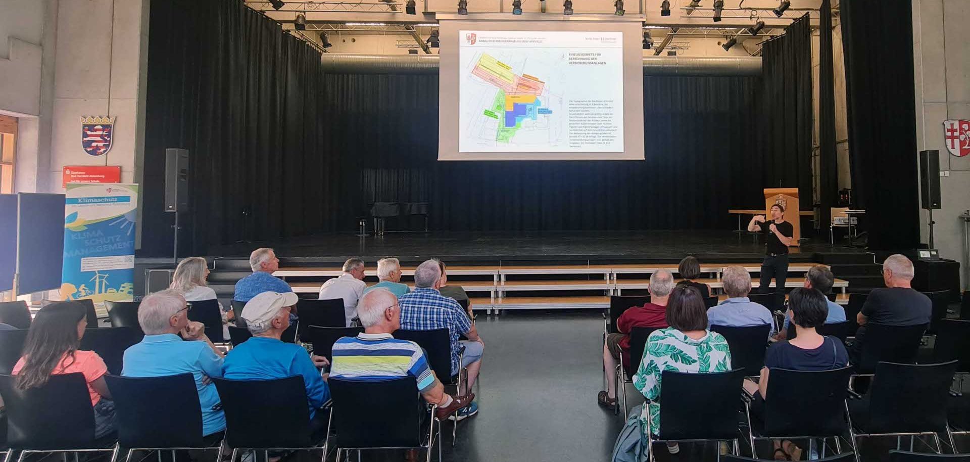 Ein Vortrag über Klimaschutz findet in einem Veranstaltungssaal statt. Etwa 20 Personen sitzen auf schwarzen Stühlen und hören einem Redner zu, der vorne auf der Bühne steht und mit einem Mikrofon spricht. Auf einer großen Leinwand ist eine Präsentationsfolie mit einer farbigen Karte und Text zu sehen. Im Hintergrund hängt ein Banner mit der Aufschrift ‚Klimaschutzmanagement‘. Der Raum wirkt modern, mit einer Bühne, Vorhängen und Wappen an den Wänden.
