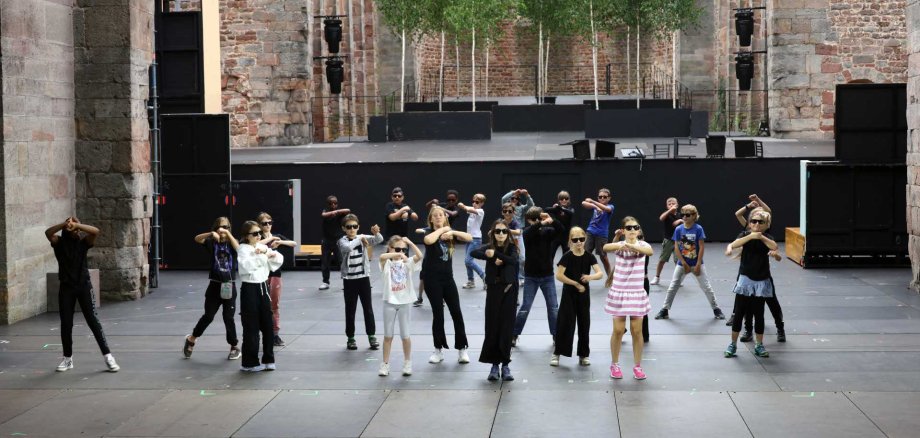 Etwa 20 Kinder tanzen synchron auf der Bühne in einer alten Klosterruine. Alle tragen Sonnenbrillen und führen eine Choreografie mit überkreuzten Armen aus. Die Bühne ist von imposanten Backsteinmauern und Säulen umgeben, im Hintergrund sind junge Birkenbäume zu sehen.