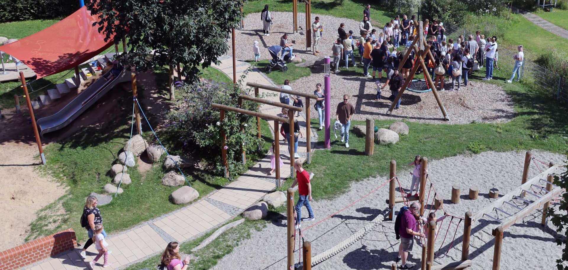 Eine große Gruppe von Menschen, überwiegend Erwachsene mit Kindern, hat sich auf einem Spielplatz bei sonnigem Wetter versammelt. Im Zentrum befindet sich eine große Nestschaukel aus Holz, die aktuell nicht genutzt wird. Die Gruppe steht dicht gedrängt auf einer Rasenfläche und dem angrenzenden mit Rindenmulch bedeckten Bereich. Viele Kinder sind von Erwachsenen begleitet. Im Hintergrund sieht man grüne Hecken, einen Gartenweg und ein weißes Gartenmöbelstück mit einem bunt dekorierten Schild. Der Spielplatz liegt in einer gepflegten, naturnahen Umgebung mit Blumen, Wiese und Bäumen.