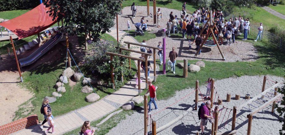 Eine große Gruppe von Menschen, überwiegend Erwachsene mit Kindern, hat sich auf einem Spielplatz bei sonnigem Wetter versammelt. Im Zentrum befindet sich eine große Nestschaukel aus Holz, die aktuell nicht genutzt wird. Die Gruppe steht dicht gedrängt auf einer Rasenfläche und dem angrenzenden mit Rindenmulch bedeckten Bereich. Viele Kinder sind von Erwachsenen begleitet. Im Hintergrund sieht man grüne Hecken, einen Gartenweg und ein weißes Gartenmöbelstück mit einem bunt dekorierten Schild. Der Spielplatz liegt in einer gepflegten, naturnahen Umgebung mit Blumen, Wiese und Bäumen.