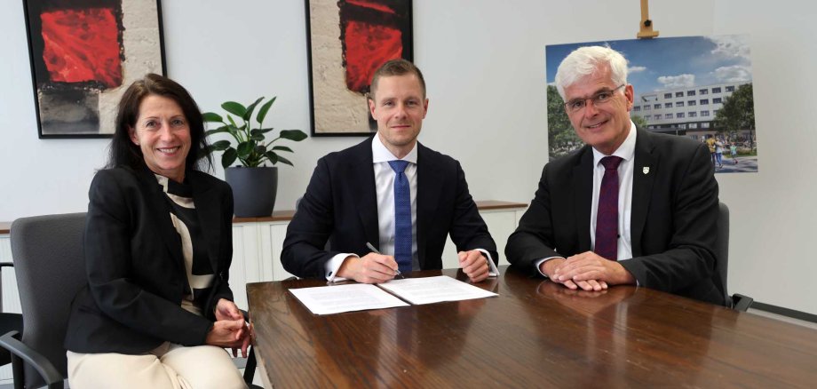 Drei Personen sitzen an einem Konferenztisch in einem modernen Büro. In der Mitte sitzt ein Mann im Anzug mit blauer Krawatte, der ein Dokument unterschreibt. Links neben ihm sitzt eine Frau in heller Hose und schwarzem Blazer, sie lächelt in die Kamera. Rechts sitzt ein älterer Mann mit weißem Haar, Brille und dunklem Anzug. Im Hintergrund sind zwei abstrakte Bilder und eine große Pflanze zu sehen. Zudem steht auf einer Staffelei ein Poster mit einem Gebäudeentwurf. Die Szene wirkt offiziell und professionell – vermutlich ein Vertragsabschluss oder ein Treffen mit politischem oder wirtschaftlichem Kontext.