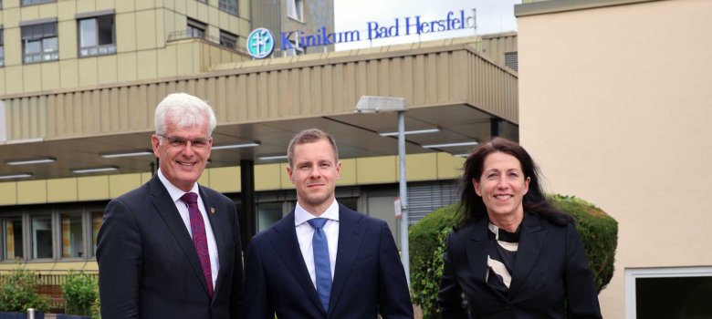 Drei Personen stehen im Freien vor dem Gebäude des „Klinikum Bad Hersfeld“. In der Mitte steht ein Mann im dunkelblauen Anzug mit hellblauer Krawatte, rechts neben ihm eine Frau in heller Hose und dunklem Blazer, links ein älterer Mann mit weißem Haar, Brille und ebenfalls im Anzug mit heller Hose und roter Krawatte. Alle drei lächeln in die Kamera. Im Hintergrund ist gut sichtbar das Klinikgebäude mit dem Schriftzug „Klinikum Bad Hersfeld“ sowie Teile der Außenanlage mit Pflanzen und Pflastersteinen. Die Szene vermittelt einen offiziellen Anlass, vermutlich im Zusammenhang mit dem Krankenhaus.