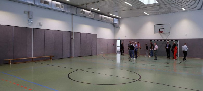 Personen stehen verteilt in der Turnhalle, im Hintergrund ist ein Basketballkorb und ein Tor zu sehen.