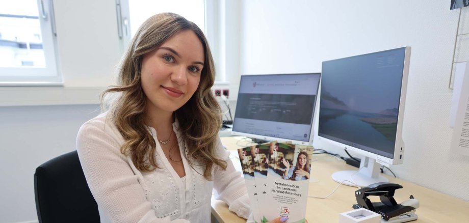 Eine junge Frau sitzt an einem Schreibtisch mit zwei Monitoren und hält mehrere Infoflyer mit dem Titel „Verfahrenslotsin im Landkreis Hersfeld-Rotenburg“ in der Hand. Sie blickt freundlich in die Kamera.