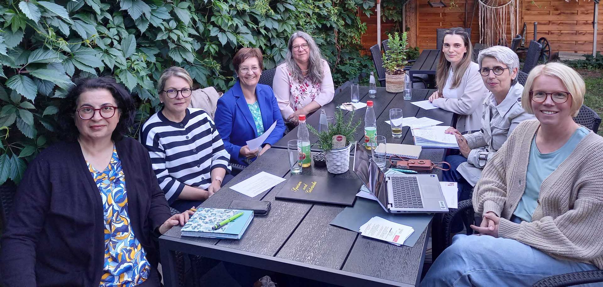 Eine Gruppe von sieben Frauen sitzt bei einem Treffen im Freien an einem langen, dunklen Gartentisch. Sie befinden sich in einem gemütlichen Garten mit viel Grün und Holzwand im Hintergrund, geschmückt mit Makramee-Dekoration. Die Frauen wirken engagiert und freundlich, einige haben Notizblöcke, Papiere und Laptops vor sich. Es scheint sich um ein informelles Arbeits- oder Netzwerktreffen zu handeln. Die Atmosphäre ist entspannt und kollegial.