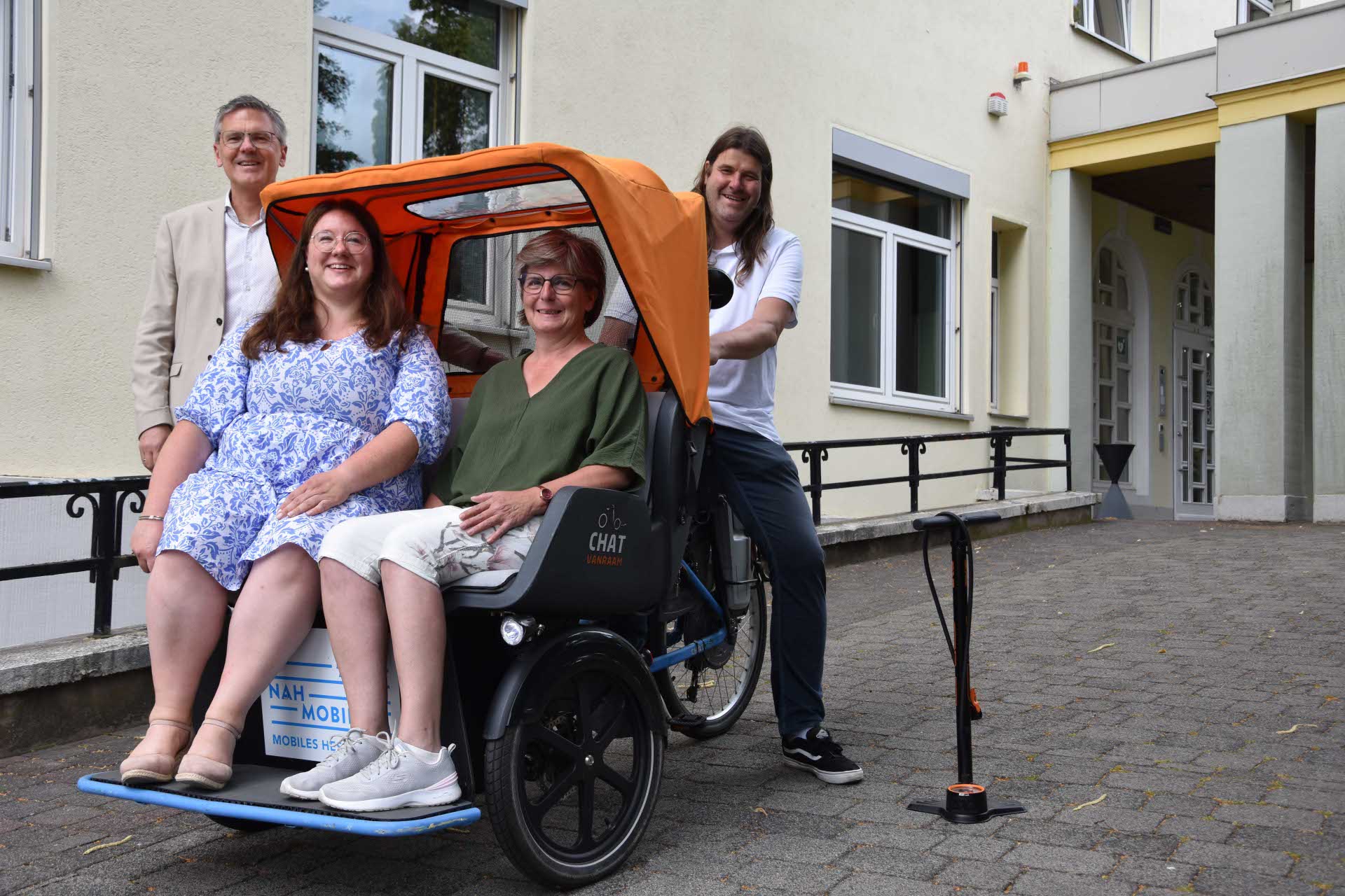 Vier Personen stehen und sitzen vor einem Gebäude neben einer Rikscha – zwei Frauen sitzen darin, ein Mann steht dahinter am Lenker, ein weiterer daneben. Auf dem Boden steht eine Fahrradpumpe.