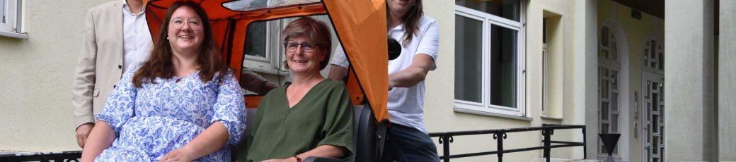 Vier Personen stehen und sitzen vor einem Gebäude neben einer Rikscha – zwei Frauen sitzen darin, ein Mann steht dahinter am Lenker, ein weiterer daneben. Auf dem Boden steht eine Fahrradpumpe.