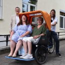 Vier Personen stehen und sitzen vor einem Gebäude neben einer Rikscha – zwei Frauen sitzen darin, ein Mann steht dahinter am Lenker, ein weiterer daneben. Auf dem Boden steht eine Fahrradpumpe.