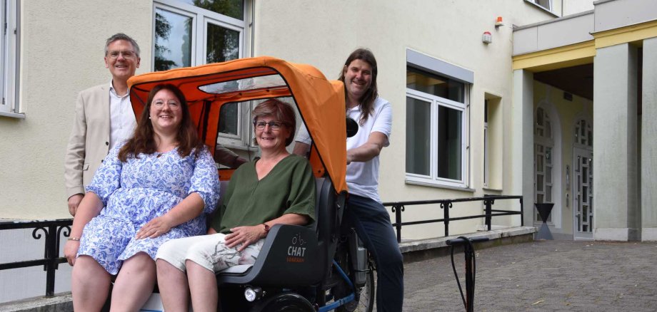 Vier Personen stehen und sitzen vor einem Gebäude neben einer Rikscha – zwei Frauen sitzen darin, ein Mann steht dahinter am Lenker, ein weiterer daneben. Auf dem Boden steht eine Fahrradpumpe.