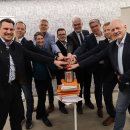 Eine Gruppe von neun Personen steht in einem modernen Bürogebäude und legt gemeinsam die Hände auf roten Buzzer mit dem hessischen Wappen. Im Hintergrund sind Banner mit den Logos von „Hessen“ und „Prosoz“ zu sehen.
