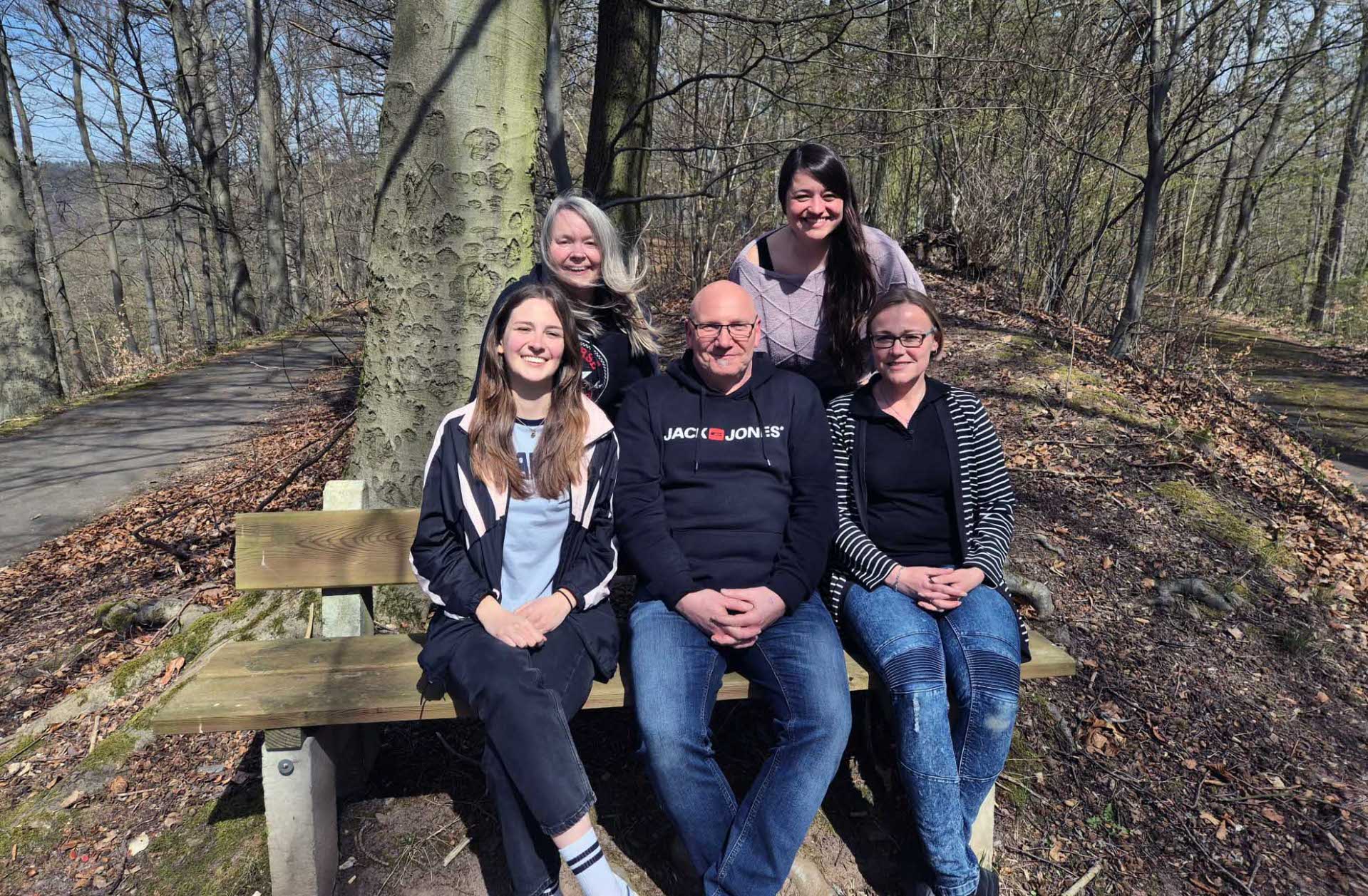 Fünf Frauen und ein Mann posieren gemeinsam für ein Gruppenfoto im Wald. Drei Personen (zwei Frauen und ein Mann) sitzen auf einer Holzbank, während zwei Frauen hinter ihnen stehen. Alle lächeln in die Kamera. Es ist ein sonniger Tag, und rundherum sind kahle Bäume ohne Laub, was auf den Frühling oder späten Herbst hindeutet. Im Hintergrund verläuft ein asphaltierter Weg durch den Wald.