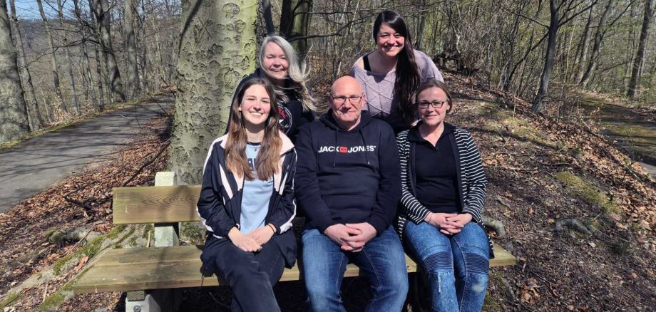Fünf Frauen und ein Mann posieren gemeinsam für ein Gruppenfoto im Wald. Drei Personen (zwei Frauen und ein Mann) sitzen auf einer Holzbank, während zwei Frauen hinter ihnen stehen. Alle lächeln in die Kamera. Es ist ein sonniger Tag, und rundherum sind kahle Bäume ohne Laub, was auf den Frühling oder späten Herbst hindeutet. Im Hintergrund verläuft ein asphaltierter Weg durch den Wald.