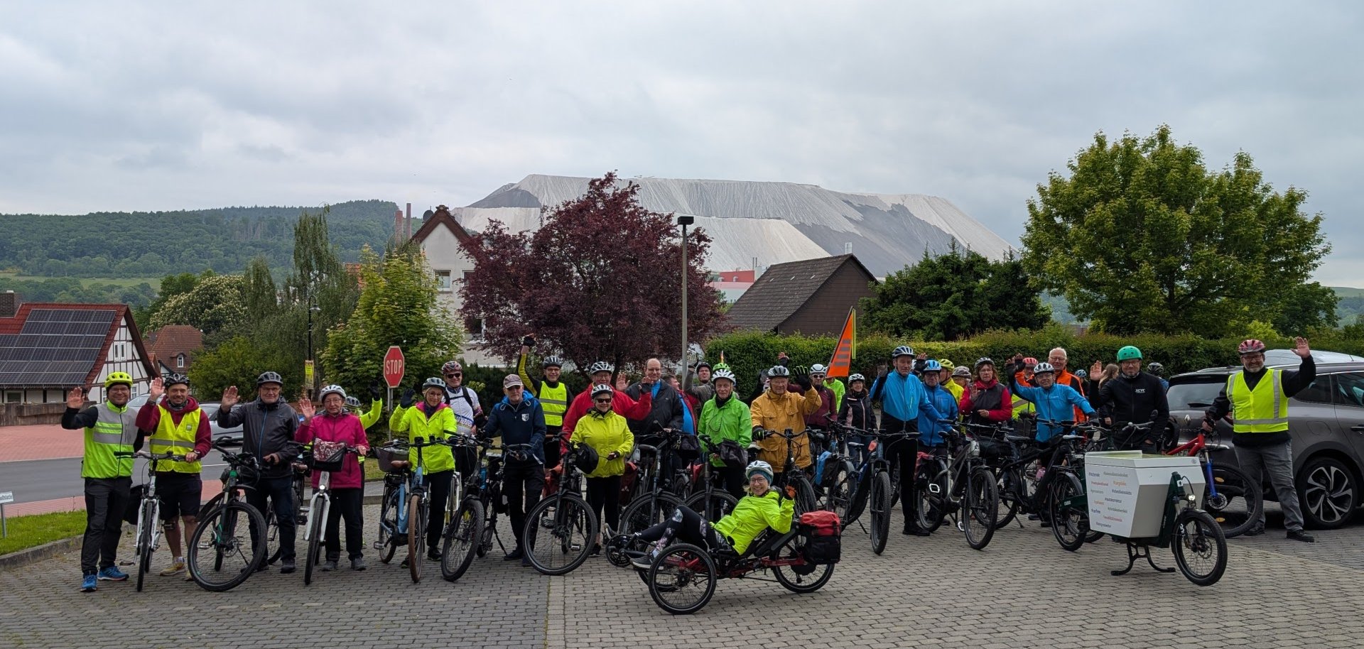Eine große Gruppe von Radfahrerinnen und Radfahrern steht mit ihren Fahrrädern auf einem gepflasterten Platz in einem Dorf. Viele tragen bunte Regenjacken und Fahrradhelme. Einige winken freundlich in die Kamera. Im Hintergrund sind Wohnhäuser mit roten Dächern und eine auffällige, große Abraumhalde eines Salzbergwerks zu sehen. Der Himmel ist bewölkt, die Stimmung wirkt dennoch fröhlich und gemeinschaftlich.