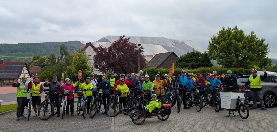 Eine große Gruppe von Radfahrerinnen und Radfahrern steht mit ihren Fahrrädern auf einem gepflasterten Platz in einem Dorf. Viele tragen bunte Regenjacken und Fahrradhelme. Einige winken freundlich in die Kamera. Im Hintergrund sind Wohnhäuser mit roten Dächern und eine auffällige, große Abraumhalde eines Salzbergwerks zu sehen. Der Himmel ist bewölkt, die Stimmung wirkt dennoch fröhlich und gemeinschaftlich.