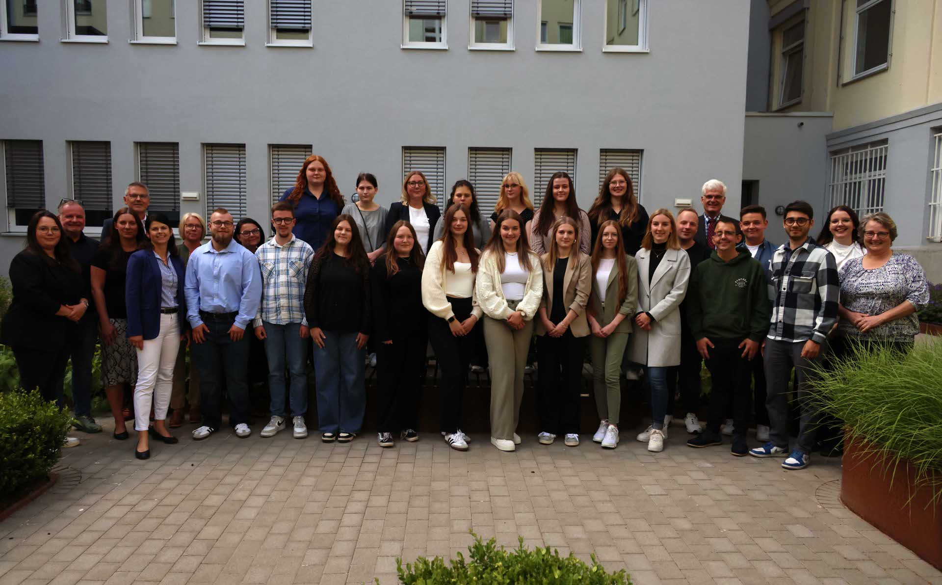 Eine große Gruppe steht in zwei Reihen für ein Gruppenfoto im Innenhof des Landratsamts. Im Hintergrund ist ein graues Gebäude mit Fenstern zu sehen. Die Personen lächeln in die Kamera.