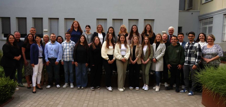 Eine große Gruppe steht in zwei Reihen für ein Gruppenfoto im Innenhof des Landratsamts. Im Hintergrund ist ein graues Gebäude mit Fenstern zu sehen. Die Personen lächeln in die Kamera.