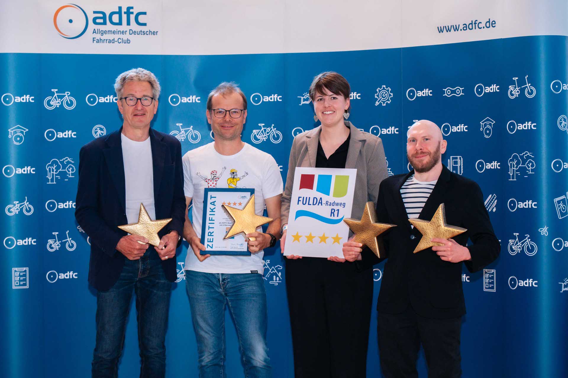 Vier Personen stehen vor einer blauen ADFC-Fotowand und halten Urkunden sowie goldene Sterne in den Händen. Eine Frau hält zudem ein Schild mit dem Logo und vier Sternen des Fulda-Radwegs R1.