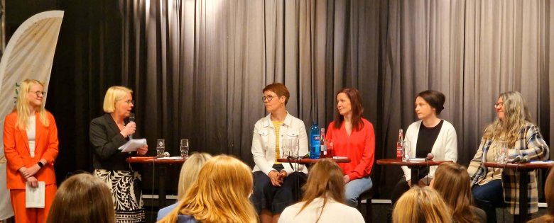 Fünf Frauen sitzen auf einer Bühne vor schwarzem Vorhang. Zwei Moderatorinnen führen ein Gespräch mit vier Landtagsabgeordneten. Auf dem Tisch vor ihnen stehen Wasserflaschen und Mikrofone, im Publikum sitzen viele Zuhörerinnen.