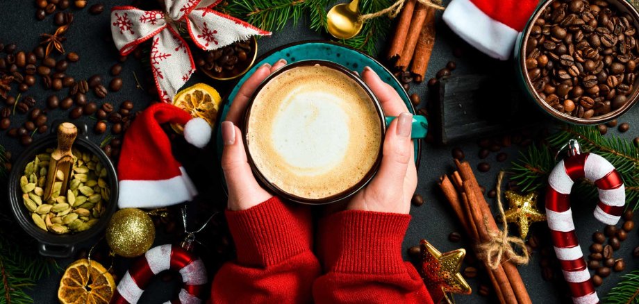 Es sind zwei Hände um eine Kaffeetasse zu sehen. Zudem liegen am Rand diverse Weihnachtsdekorationen.
