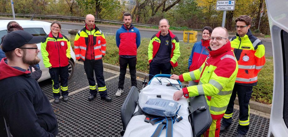 Mehrere ehrenamtliche Einsatzkräfte in Rettungsdienstkleidung stehen im Freien um eine Trage und besprechen gemeinsam ein Einsatzszenario während einer Fortbildung.