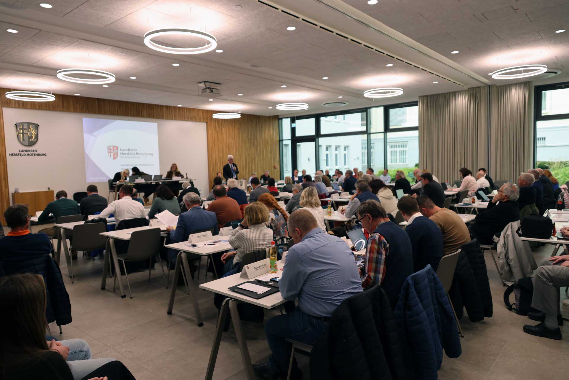 In einem Raum sind viele Menschen zu sehen. Es läuft eine Sitzung. Im Hintergrund spricht ein Mann an einem Rednerpult.