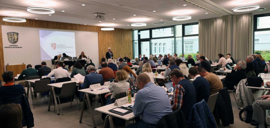 In einem Raum sind viele Menschen zu sehen. Es läuft eine Sitzung. Im Hintergrund spricht ein Mann an einem Rednerpult.