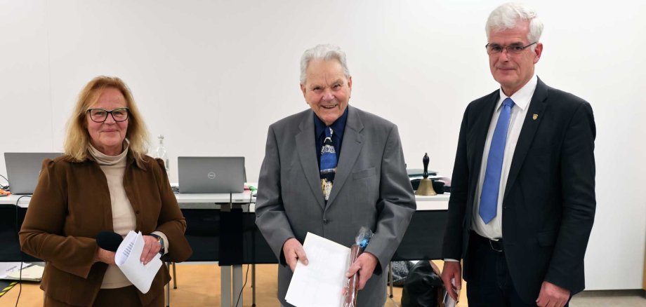 Drei ältere Personen stehen in einem Innenraum vor einem Tisch mit mehreren Laptops. Eine Frau links hält Unterlagen und ein Mikrofon, in der Mitte steht ein lächelnder Mann im Anzug mit Dokumenten in den Händen, rechts steht ein weiterer Mann im Anzug mit blauer Krawatte. Die Szene wirkt wie eine offizielle Ehrung oder eine formelle Veranstaltung.