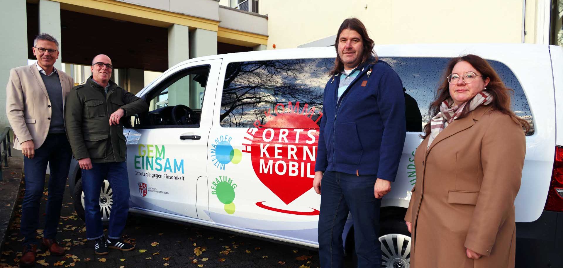 Vier Personen stehen vor einem weißen Transporter mit der Aufschrift „Hier kommt das Ortskernmobil“ und „GEMEINSAM – Strategie gegen Einsamkeit“. Das Fahrzeug ist mit bunten Logos und Schriftzügen beklebt. Die Gruppe posiert lächelnd neben dem Wagen, der auf einem mit Herbstlaub bedeckten Pflasterweg vor einem Gebäude geparkt ist.
