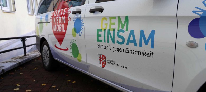 Seitenansicht eines weißen Fahrzeugs mit bunten Aufdrucken. Auf der Tür steht „GEMEINSAM – Strategie gegen Einsamkeit“ und das Logo des Landkreises Hersfeld-Rotenburg. Daneben ist ein rotes Herz mit der Aufschrift „Hier kommt das Ortskernmobil“ sowie farbige Kreise mit den Worten „Miteinander“ und „Beratung“ zu sehen. Das Auto steht auf einem gepflasterten Weg neben einem Gebäude.
