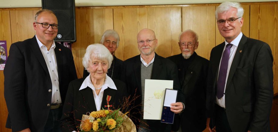Eine ältere Frau steht in der Mitte einer Gruppe von fünf Männern. Sie hält einen Blumenstrauß und trägt eine Medaille an ihrem Jackett. Einer der Männer neben ihr hält eine Urkunde und eine Schachtel mit einer weiteren Auszeichnung. Alle lächeln in die Kamera und stehen in einem Raum mit holzvertäfelter Wand – offenbar bei einer Ehrungsveranstaltung.