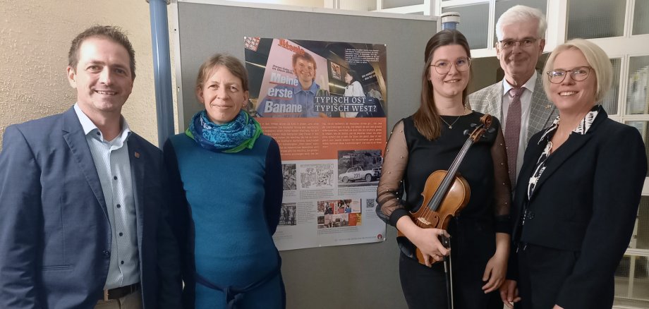 Fünf Personen stehen vor einer Stellwand mit einer Ausstellungstafel. Eine Frau in der Mitte hält eine Violine in den Händen. Die anderen vier Personen tragen überwiegend formelle Kleidung. Alle schauen in die Kamera und lächeln.