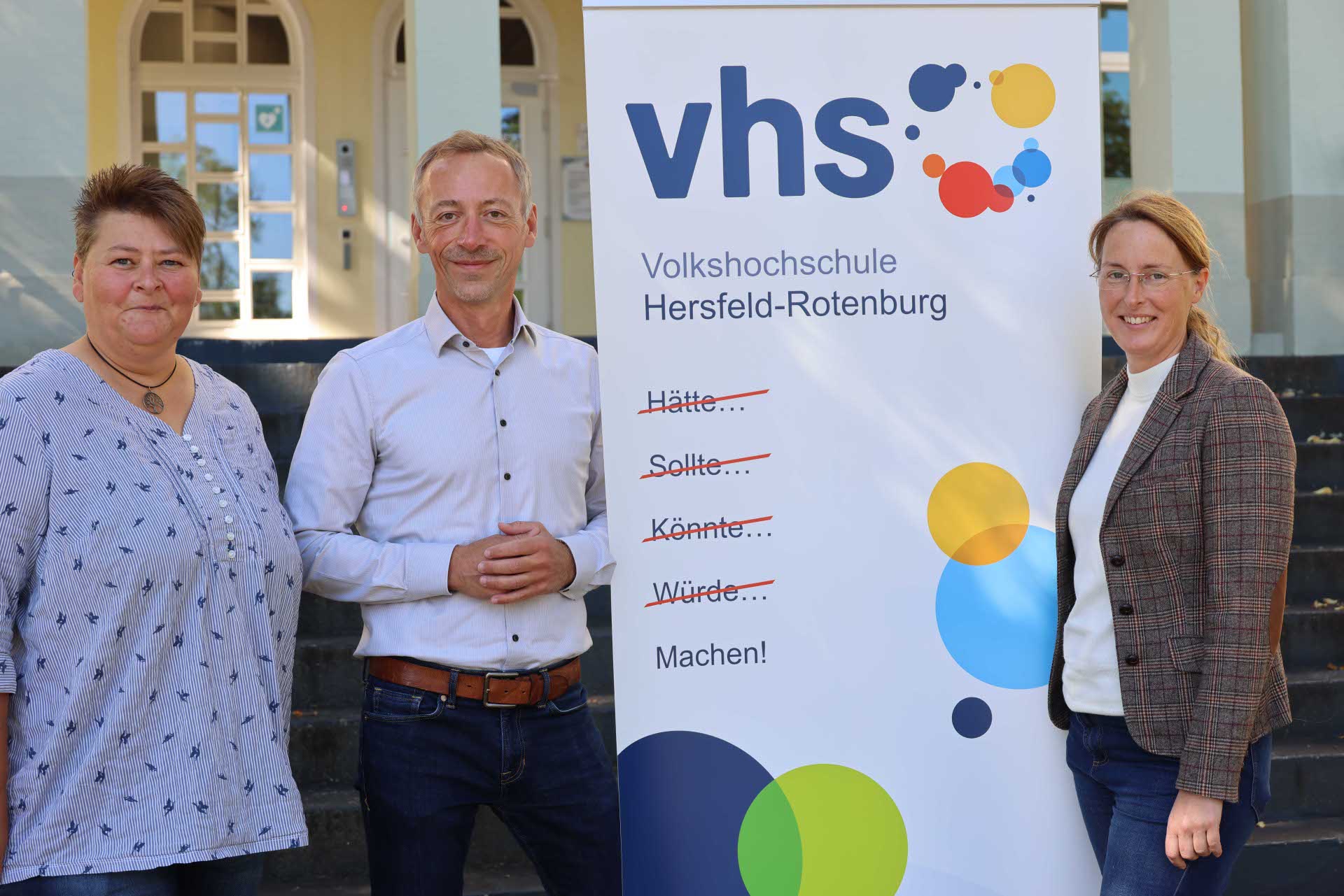 Drei Personen stehen neben einem Roll-up der Volkshochschule Hersfeld-Rotenburg, das den Slogan „Hätte… Sollte… Könnte… Würde… Machen!“ zeigt.