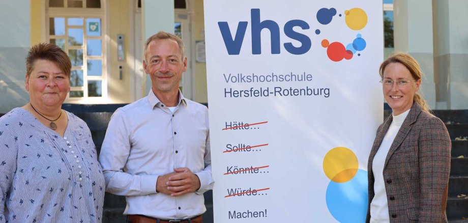 Drei Personen stehen neben einem Roll-up der Volkshochschule Hersfeld-Rotenburg, das den Slogan „Hätte… Sollte… Könnte… Würde… Machen!“ zeigt.