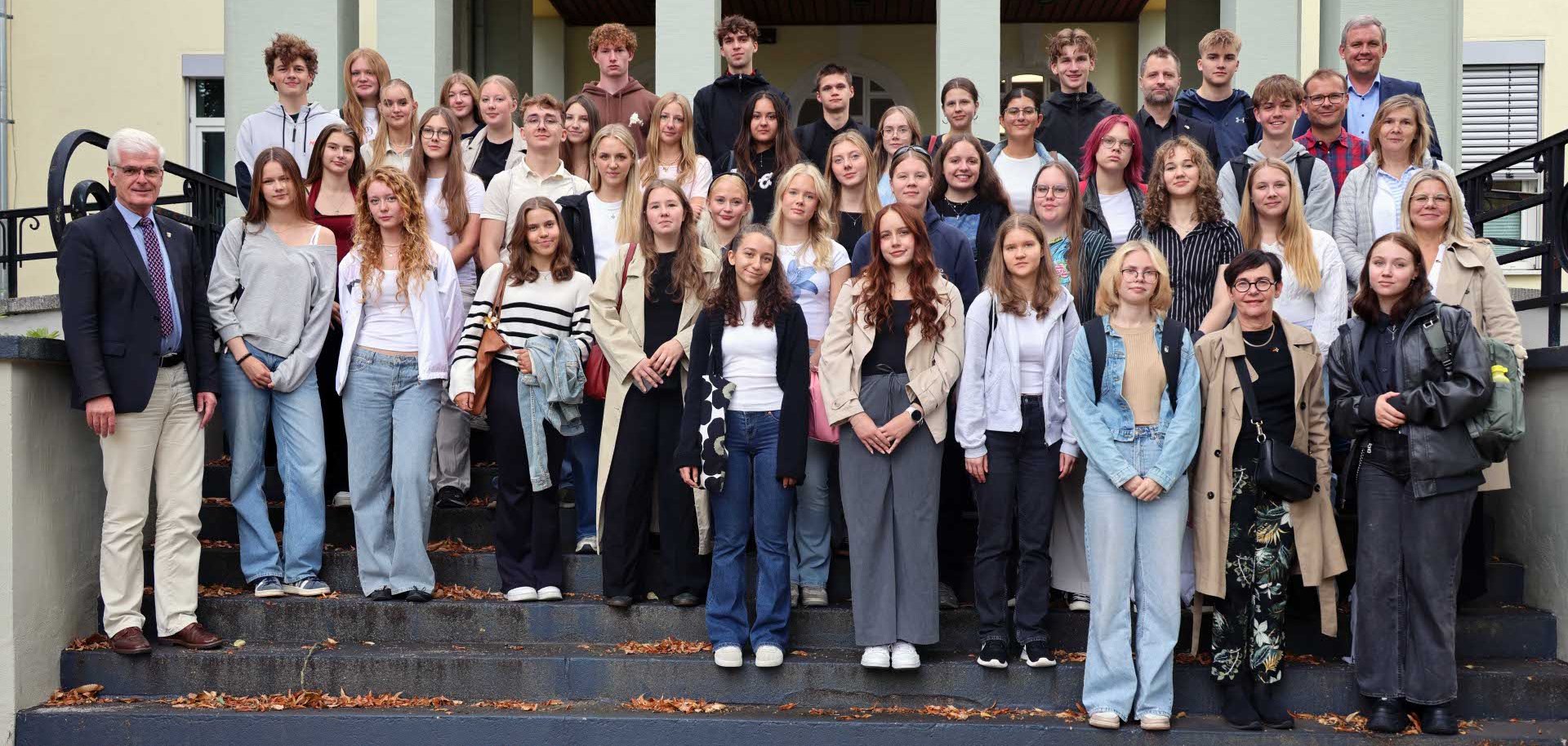 Eine große Gruppe von Schülerinnen, Schülern und Erwachsenen steht für ein gemeinsames Foto auf den Treppen vor einem Gebäude. Die Jugendlichen befinden sich in der Mitte, während einige Erwachsene am Rand und in der hinteren Reihe zu sehen sind. Alle blicken in die Kamera, viele lächeln. Im Hintergrund ist der Eingangsbereich des Gebäudes mit Säulen und einem Vordach zu erkennen.