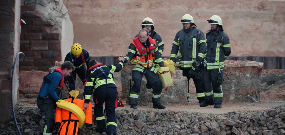 Mehrere Feuerwehrleute und Rettungskräfte bergen gemeinsam eine in einer orangefarbenen Rettungspuppe dargestellte Person aus dem Wasser. Einige Einsatzkräfte tragen Schwimmwesten und Helme, andere Feuerwehrschutzkleidung. Im Vordergrund sind große weiße Sandsäcke gestapelt, im Hintergrund eine Mauer und Geröll.