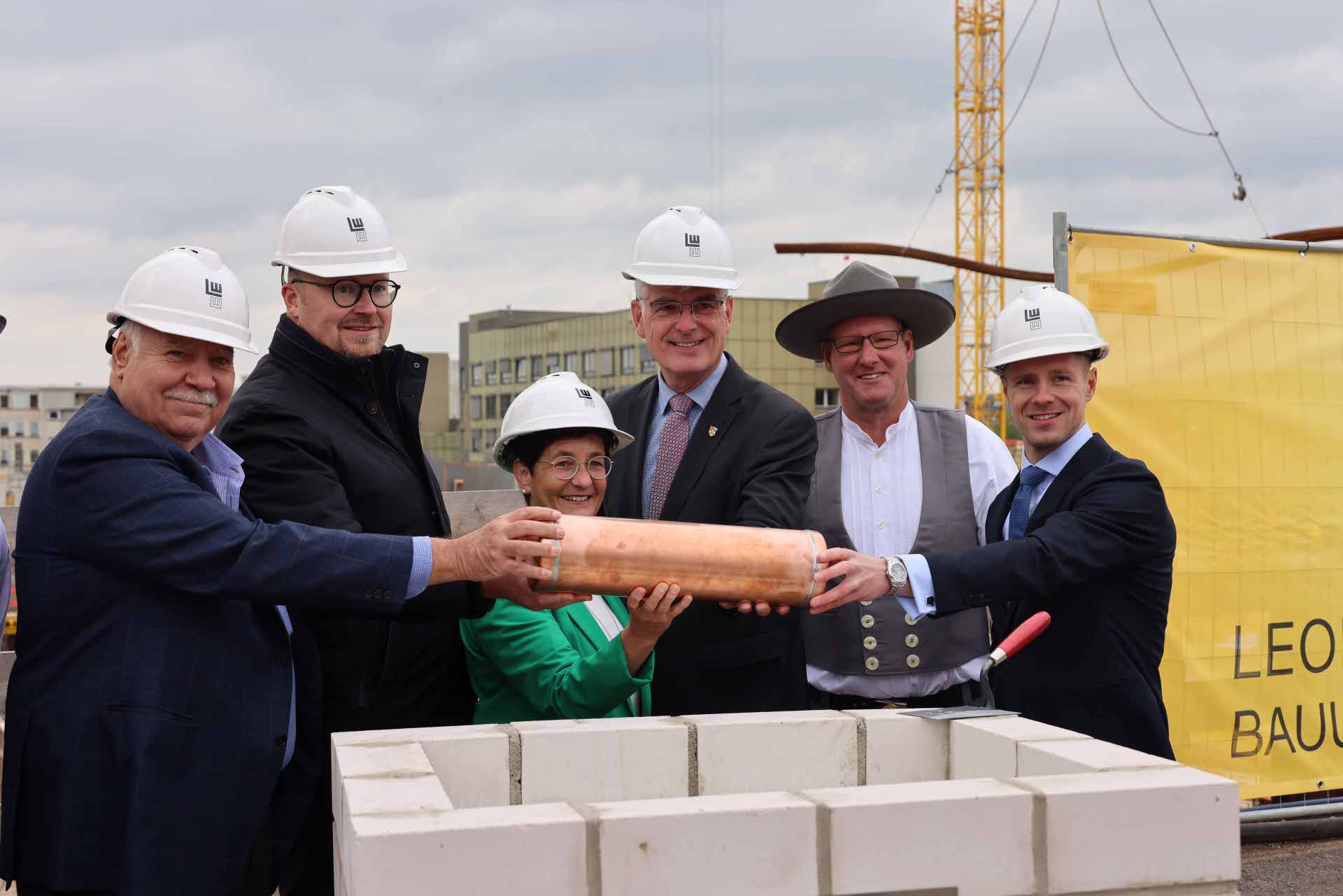 Fünf Männer und eine Frau halten eine Zeitkapsel und stehen hinter dem Grundstein. Im Hintergrund sieht man das Klinikum Bad Hersfeld und einen Kran.
