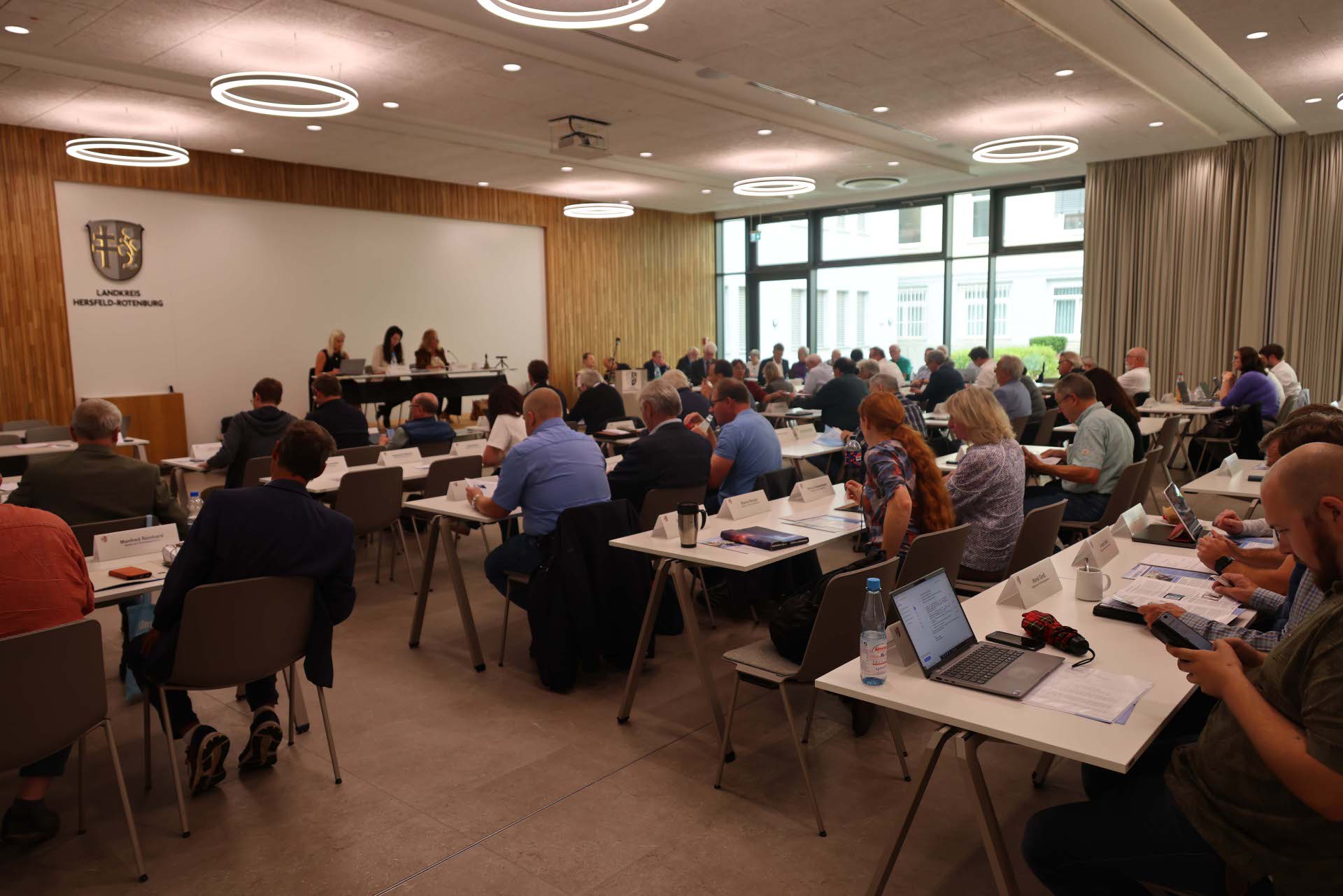 Großer Sitzungssaal im Landratsamt Hersfeld-Rotenburg. Zahlreiche Personen sitzen an Tischen, nehmen an einer Sitzung teil und arbeiten mit Unterlagen, Laptops und Tablets. Vorne sitzen drei Personen am Podium. Im Hintergrund das Logo des Landkreises an der Wand.