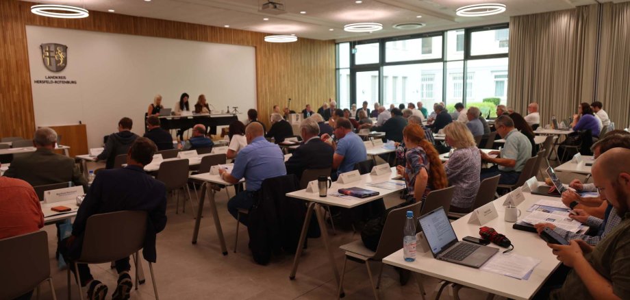 Großer Sitzungssaal im Landratsamt Hersfeld-Rotenburg. Zahlreiche Personen sitzen an Tischen, nehmen an einer Sitzung teil und arbeiten mit Unterlagen, Laptops und Tablets. Vorne sitzen drei Personen am Podium. Im Hintergrund das Logo des Landkreises an der Wand.