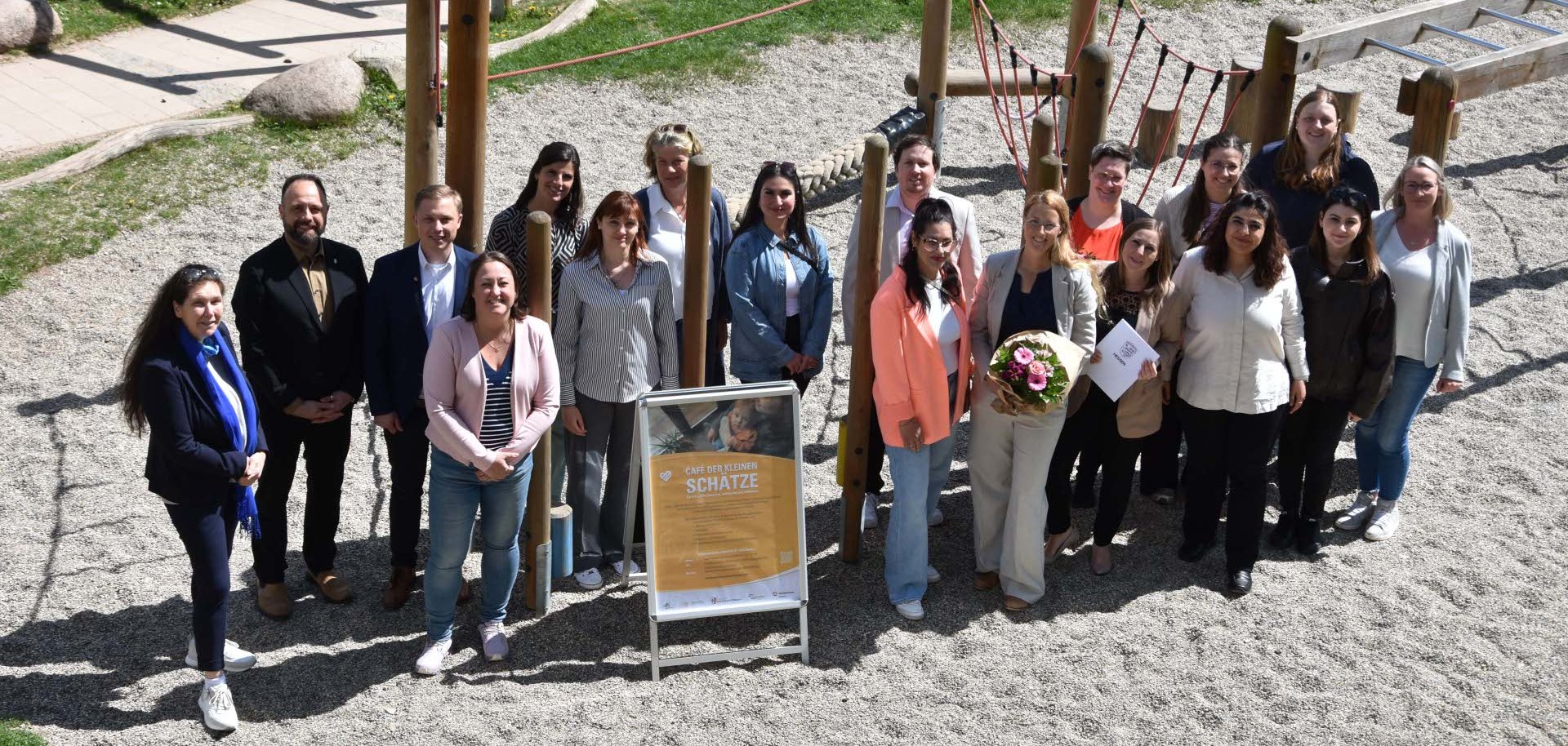 Eine Gruppe von mehreren Erwachsenen steht auf einem Kinderspielplatz. Sie schauen alle in die Kamera. Zwei Personen haben einen Blumenstrauß in der Hand, eine Person hält eine Urkunde. 