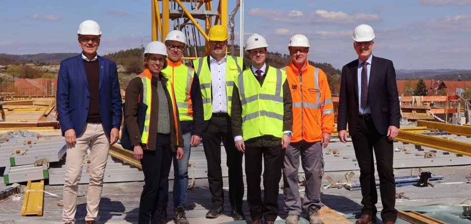 Gruppe von sieben Personen auf einer Baustelle, alle mit Schutzhelmen; einige tragen Warnwesten, andere Anzüge. Sie stehen vor einem gelben Baukran auf einer im Bau befindlichen Fläche mit Baumaterialien, bei sonnigem Wetter und ländlicher Umgebung im Hintergrund.