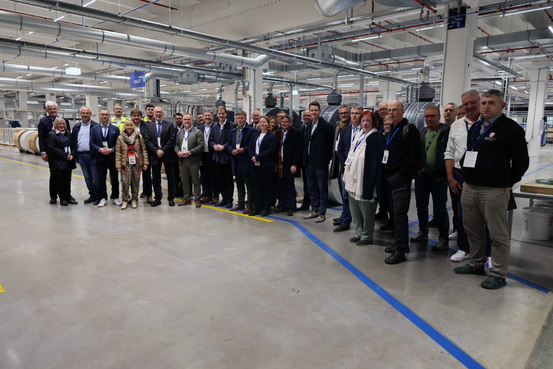 Eine größere Gruppe von Menschen steht nebeneinander in einer Produktionshalle und blickt in die Kamera. Die meisten tragen Business- oder Smart-Casual-Kleidung sowie Namensschilder. Im Hintergrund sind industrielle Maschinen, Rohre und große Materialrollen zu sehen. Die Umgebung wirkt sauber, modern und technisch geprägt.