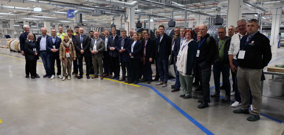 Eine größere Gruppe von Menschen steht nebeneinander in einer Produktionshalle und blickt in die Kamera. Die meisten tragen Business- oder Smart-Casual-Kleidung sowie Namensschilder. Im Hintergrund sind industrielle Maschinen, Rohre und große Materialrollen zu sehen. Die Umgebung wirkt sauber, modern und technisch geprägt.